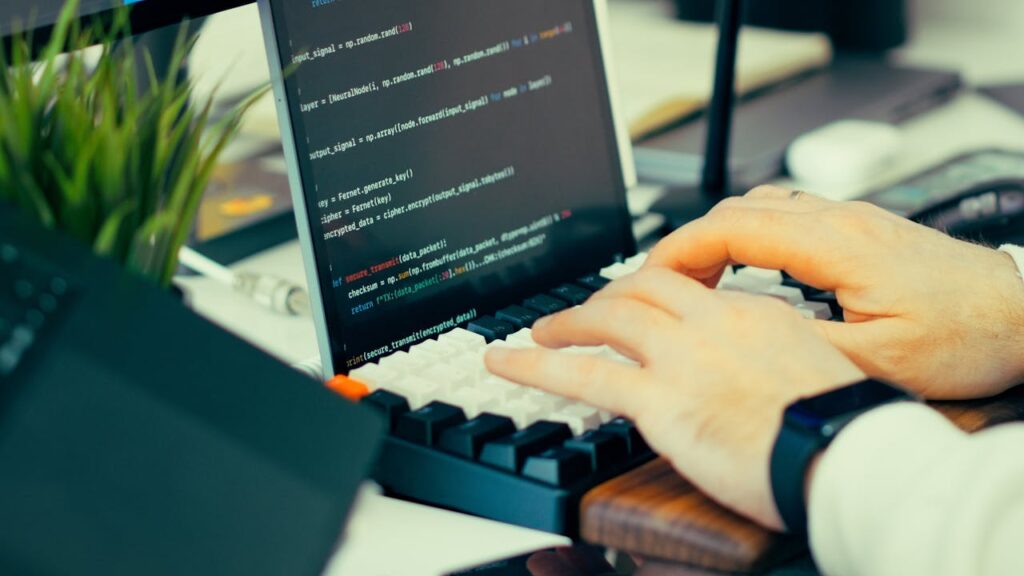 pexels photo 36497969 Close-up view of a developer typing code on a keyboard with a computer screen showing scripts.